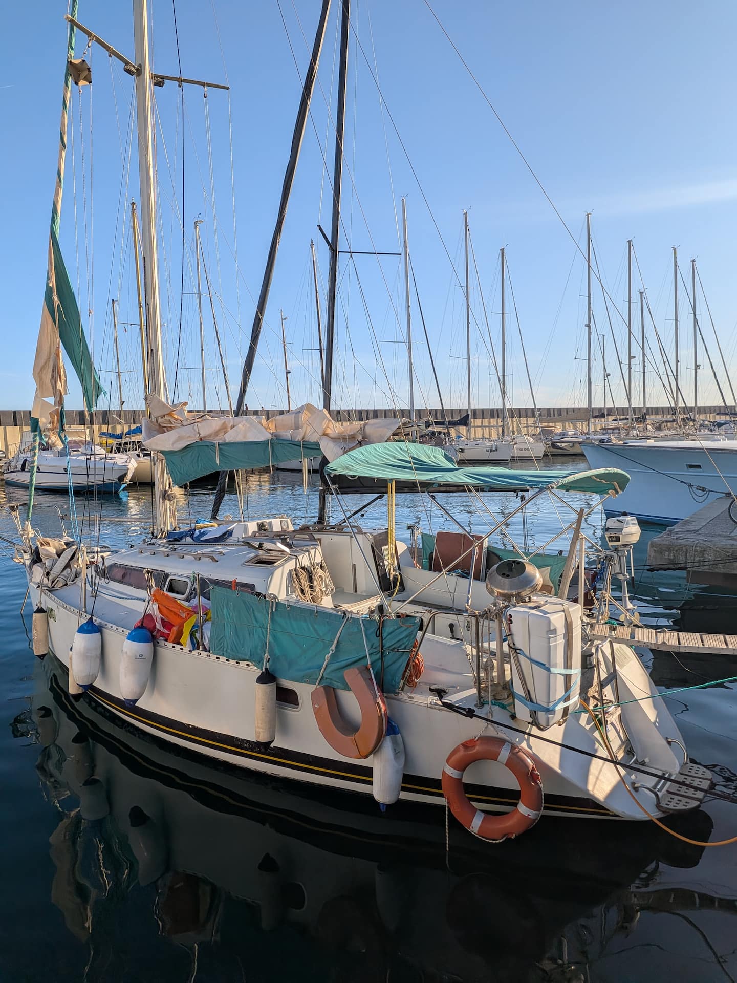 The Varazze sailboat seen from the pontoon on its berth, teal covers and lifebuoy visible, other yachts and the marina behind it.