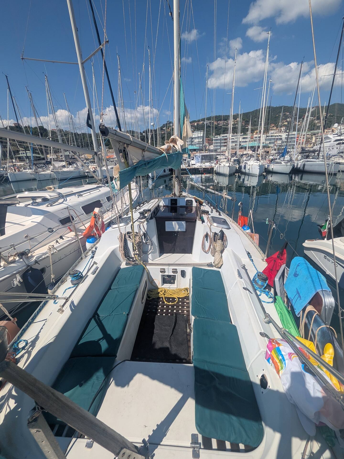 Looking forward along the deck of the Varazze sailboat from the cockpit, teal sail cover and coachroof in the foreground, masts of the marina and the Ligurian hills beyond.