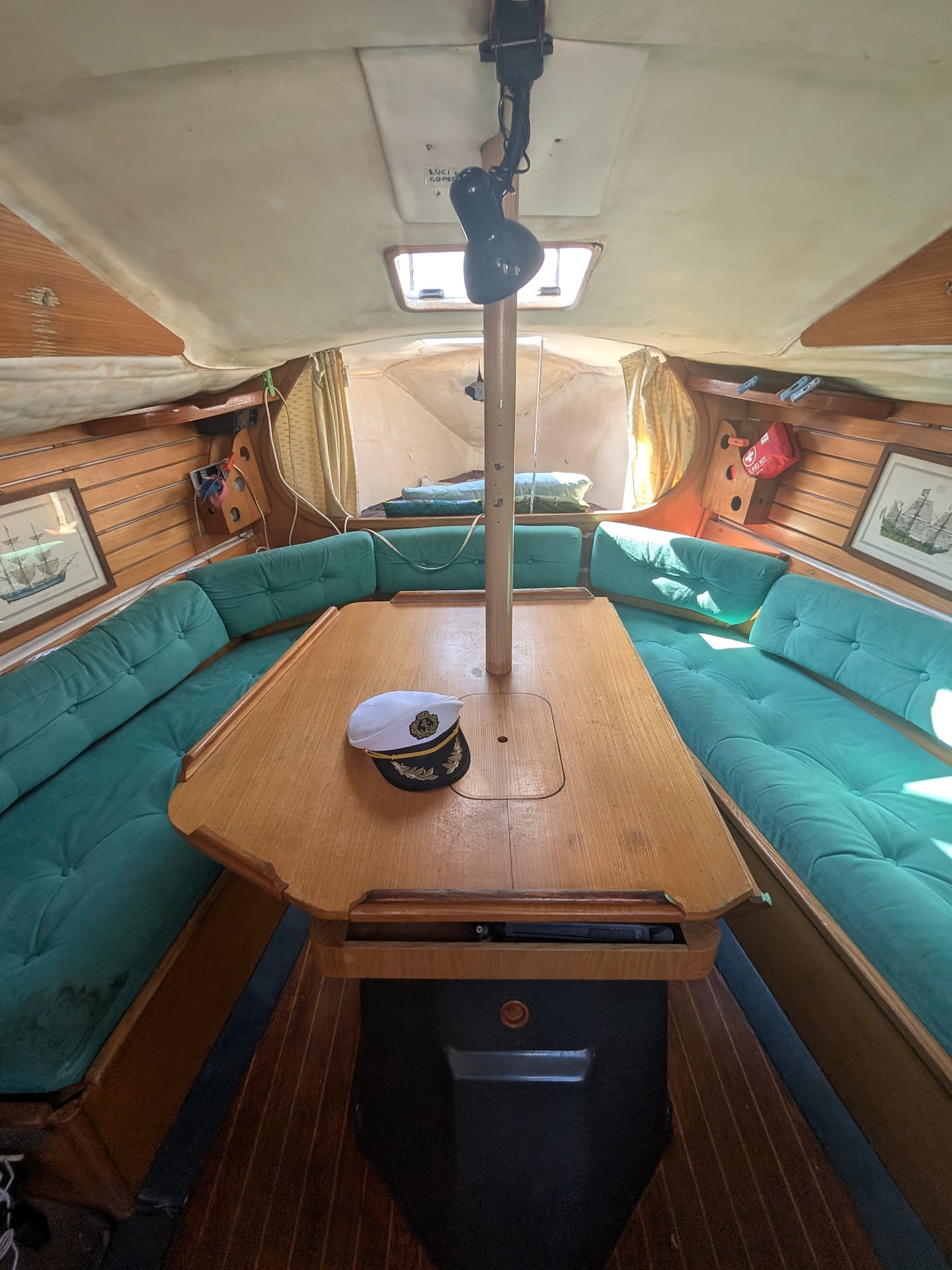 The saloon of the Varazze sailboat: a horseshoe of teal buttoned cushions around a varnished timber table, with the mast passing through the middle.
